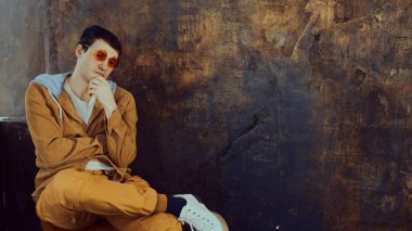 Gözlüklü genç yakışıklı adam sandalyede oturuyor ve başka tarafa bakıyor. Pejmürde kahverengi duvarın arkasında şık bir erkek.