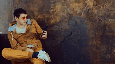 Parlak gözlüklü şık adam sandalyede oturuyor ve eski püskü kahverengi duvarın yanında cep telefonuna bakıyor. İlgilenen erkek kahverengi arkaplan üzerinde akıllı telefon kullanıyor ve metin için boşluk var