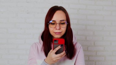 Pembe kapüşonlu uzun saçlı, gözlüklü, cep telefonuyla mesajlaşan beyaz tuğla duvara yaslanmış genç bir kadın.