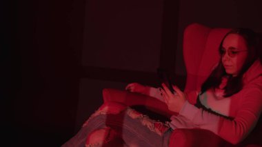 Koltukta akıllı telefondan mesaj atan bir kadın. Rahat koltukta dinlenen rahat giysili ve gözlüklü sakin genç bayanın yan görüntüsü ve kırmızı ışıklı karanlık odada akıllı telefon kullanımı..