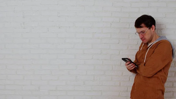 Młody Człowiek Okularach Przeglądający Telefon Komórkowy Białej Ceglanej Ścianie Stylowy — Zdjęcie stockowe
