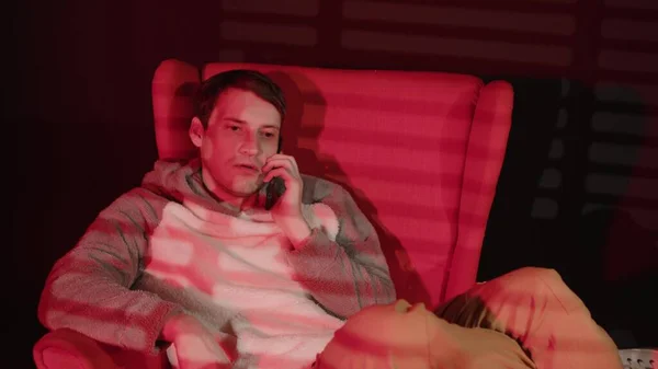 若い男は 赤い光と影のブラインドが落ちる暗い部屋のアームチェアに座って スマートフォンで話します 忙しい男性は携帯電話で話す — ストック写真