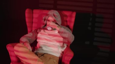 Genç adam kahve içer, sandalyede oturur ve jaluzinin gölgeleriyle altını çizer. Rahat erkek sıcak içkinin tadını çıkarıyor ve düşen kırmızı ışıkla karanlık odada dinleniyor..