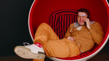 Cep telefonlu genç adam siyah arka planda top sandalyesinde oturuyor. Erkek akıllı telefonda konuşuyor ve modern koltukta dinleniyor.