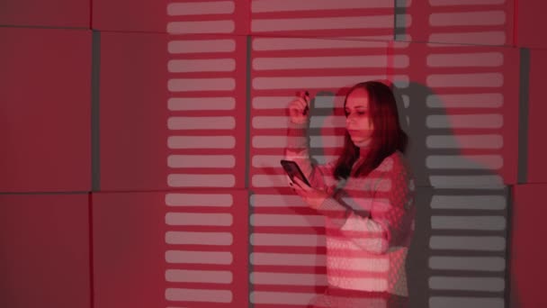 Mujer joven con teléfono móvil de pie cerca de la pared destacando con sombras de jalousie. Mujer seria usando teléfono inteligente, apoyado contra la pared en habitación oscura con la caída de la luz roja. — Vídeo de stock