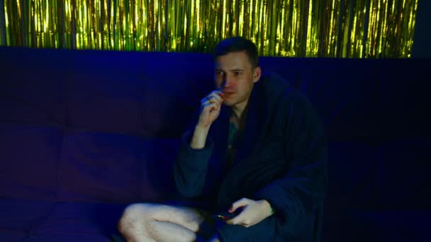 Mladý muž v županu, sedící na pohovce v tmavé místnosti s mihotavým světlem. Bezstarostný muž jíst sušenky a dívat se na televizi, v pozdních večerních hodinách. — Stock video