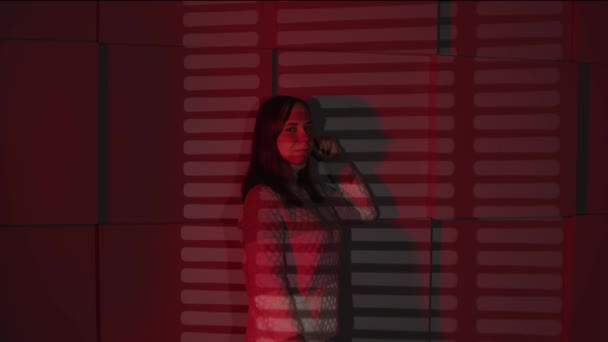 Ung kvinna står nära väggen och markerar med skuggor av jalousie. Positiv kvinnlig leende och tittar på kameran, lutar sig mot väggen i mörkt rum med fallande rött ljus. — Stockvideo
