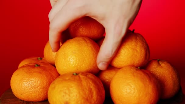 Крупним планом руки чоловіка беруть мандарин, купа мандаринів на червоному тлі — стокове відео