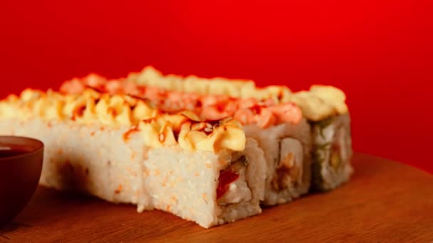 Sushi rola perto de molho de soja. Conjunto de saborosos rolos de sushi com queijo coberto com molho servido em mesa de madeira contra fundo vermelho na sala de luz — Vídeo de Stock