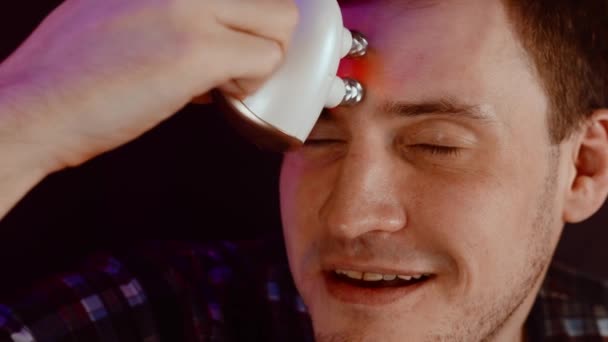 Молодой человек использует 3D массажер для лица. Расслабленный самец с закрытыми глазами с помощью электромассажера на лбу против морщин. Концепция ухода за мужчинами и косметические процедуры на дому. Закрыть. — стоковое видео
