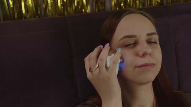 Una mujer con piel problemática usando masajeador eléctrico facial. Mujer que usa masajeador eléctrico en la cara mientras se relaja en el sofá y hace el procedimiento de belleza en casa. Concepto de control del acné — Vídeos de Stock