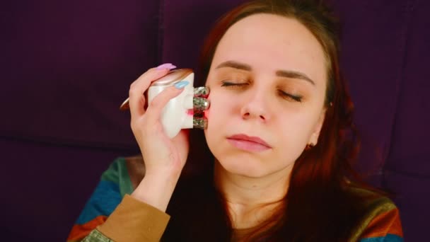 Uvolněná žena používající elektrický masážní přístroj. Žena používající elektrický masážní přístroj na obličej při relaxaci na pohovce a při kosmetické proceduře doma. Koncept salonu krásy doma — Stock video