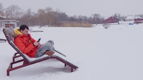 Muž v zimním oranžovém saku odpočívá a v zimě si prohlíží smartphone na osamělé lehátku u zamrzlého rybníka za sněhového počasí. Lehátko v zimě na sněhu na pláži — Stock video