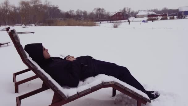 Een man met een bril rust in een zonnebank in de winter, in de buurt van een bevroren vijver in de winter bij sneeuwweer en kijkt in de verte. Een zonnebank in de winter op de sneeuw op het strand — Stockvideo