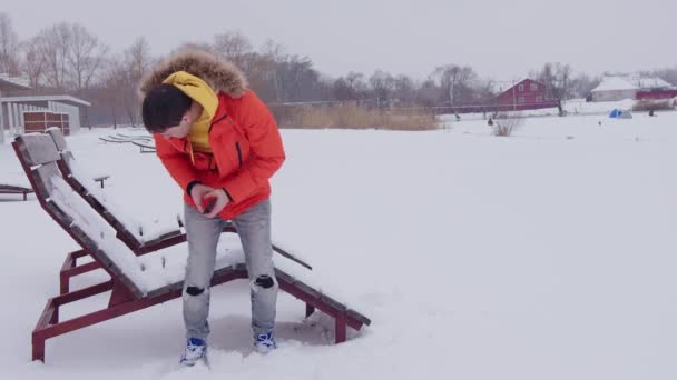 Un hombre vestido con una chaqueta naranja de invierno está descansando y viendo un teléfono inteligente en una chaise longue en invierno, cerca de un estanque congelado en invierno con clima nevado. Una tumbona en invierno en la nieve en la playa — Vídeos de Stock