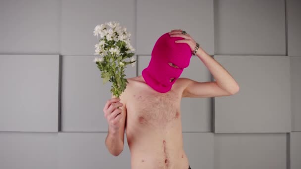Anonimowy mężczyzna w kominiarce z kwiatami. Nierozpoznawalny niebezpieczny samiec z nagim tułowiem noszący różową kominiarkę z białymi kwiatami w ręku stojący w pobliżu szarej teksturowanej ściany geometrycznej — Wideo stockowe