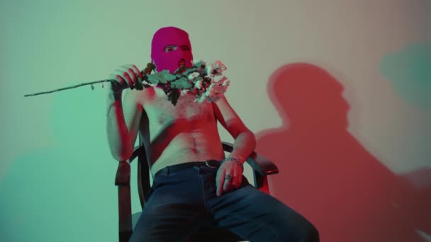 Анонимный мужчина без рубашки в балаклаве сидит с цветами. Неузнаваемый опасный мужчина с обнаженным туловищем в розовой балаклаве, сидящий в кресле у стены с букетом белых цветов в руке — стоковое видео