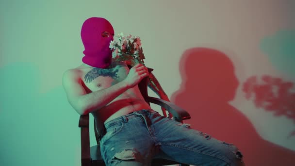 Anonyme homme torse nu en cagoule assis avec des fleurs. Homme dangereux méconnaissable avec torse nu en cagoule rose assis sur une chaise près du mur avec bouquet de fleurs blanches à la main — Video