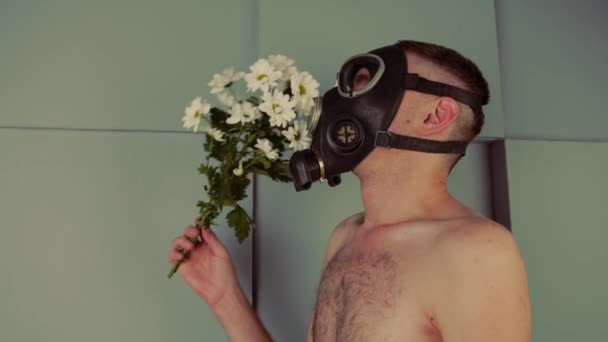 Muž bez trička v plynové masce, vonící květinami. Boční pohled na nerozpoznatelného samce s nahým trupem v černé plynové masce vonící kyticí čerstvých bílých květin u šedé stěny — Stock video