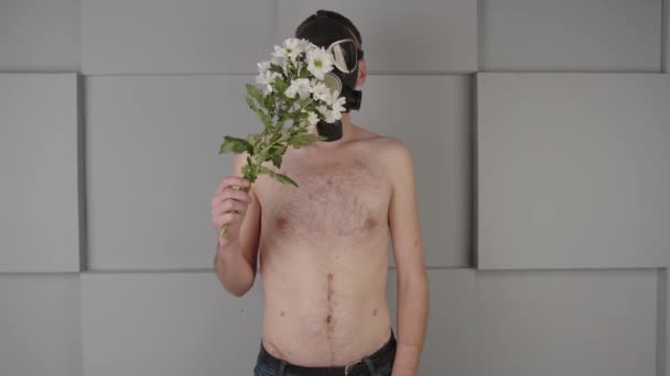 Sans visage, torse nu, masque à gaz, sentant les fleurs. Homme méconnaissable avec torse nu en masque à gaz noir bouquet odorant de fleurs blanches fraîches près du mur gris. — Video