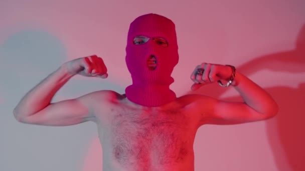Anonymous shirtless man in balaclava showing biceps. Unrecognizable male with naked torso wearing pink balaclava showing biceps and looking at camera while standing near wall with shadow — Stock Video