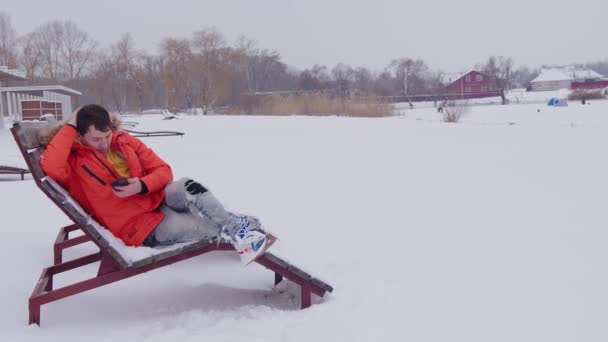 Een man gekleed in een winter oranje jas is rusten en het bekijken van een smartphone op een chaise longue in de winter, in de buurt van een bevroren vijver in de winter bij besneeuwd weer. Een zonnebank in de winter op de sneeuw op het strand — Stockvideo