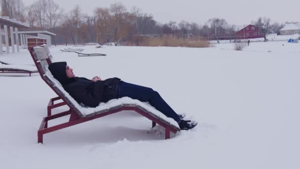 Un uomo che indossa occhiali riposa in un lettino in inverno, vicino a uno stagno ghiacciato in inverno con tempo nevoso e guarda in lontananza. Un lettino in inverno sulla neve sulla spiaggia — Video Stock