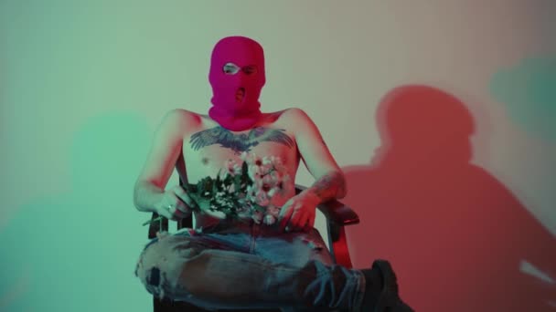Anonymní muž bez trička v balaklávě, sedí s květinami. Nerozpoznatelný nebezpečný pes s nahým trupem v růžové balaklávě sedí v křesle u zdi s kyticí bílých květin v ruce — Stock video