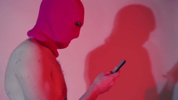 バラクラバの匿名の無名の男がスマートフォンを閲覧。側ビューの危険な認識不能な男性とともに裸の胴でbalaclavaテキストメッセージングで現代の携帯電話でネオン光の中に立って — ストック動画