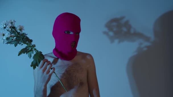 Anonymous shirtless man in balaclava with flowers. Unrecognizable dangerous male with naked torso wearing pink balaclava with bouquet of chamomile in hand standing near wall in dark room. — Stock Video