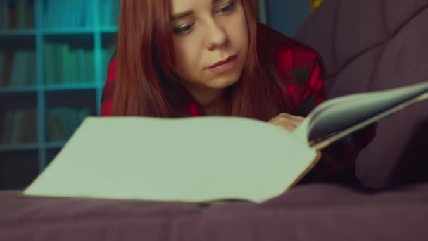 Прекрасный студент читает большую книгу. Задумчивая молодая женщина, лежащая на диване с книгой и читающая в темной комнате вечером — стоковое видео