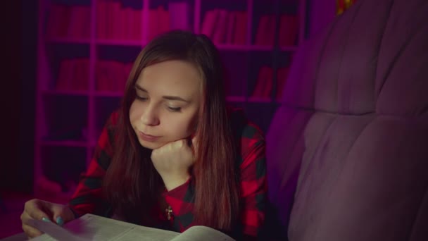 Uma bela aluna a ler um grande livro. Uma jovem pensativa deitada no sofá com um livro e lendo em um quarto escuro à noite — Vídeo de Stock
