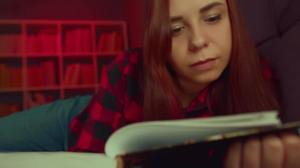 Una hermosa estudiante leyendo un gran libro. Una joven reflexiva tumbada en el sofá con un libro y leyendo en una habitación oscura por la noche — Vídeos de Stock