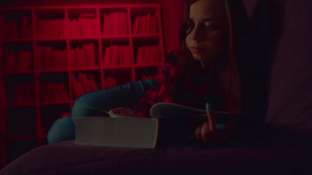 Een mooie student die een groot boek leest. Een attente jonge vrouw liggend op de bank met een boek en lezen in een donkere kamer in de avond — Stockvideo