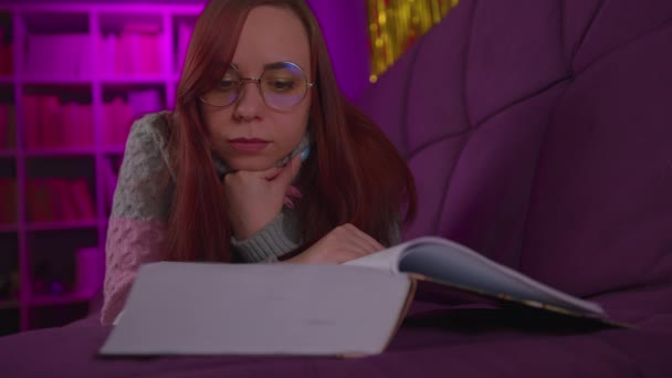Um estudante bonito com óculos lendo um grande livro. Uma jovem pensativa em uma camisola deitada no sofá com um livro e lendo à noite em um quarto escuro — Vídeo de Stock