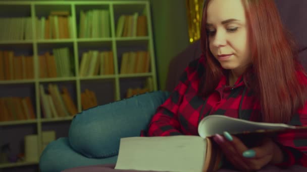 Una hermosa estudiante leyendo un gran libro. Una joven reflexiva tumbada en el sofá con un libro y leyendo en una habitación oscura por la noche — Vídeos de Stock