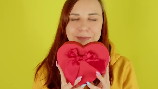 Mujer joven con caja de regalo roja en forma de corazón sobre fondo amarillo. Bonita hembra sostiene regalo romántico, mirando a la cámara y sonriendo. Concepto de presente en el día de San Valentín y el Día Internacional de la Mujer. — Vídeo de stock