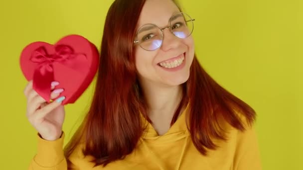 Mujer joven en gafas con caja de regalo roja en forma de corazón sobre fondo amarillo. Feliz mujer sacudiendo regalo romántico y sonriendo. Concepto de presente en el día de San Valentín y el Día Internacional de la Mujer. — Vídeos de Stock