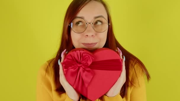 Жінка в окулярах з червоною подарунковою коробкою у вигляді серця на жовтому тлі. Мрійлива самка тримає романтичний подарунок, посміхається і дивиться геть. Концепція сьогодення в день валентинки та міжнародний жіночий день . — стокове відео