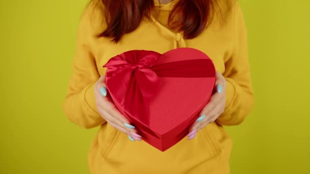 Parte del cuerpo de la mujer con caja de regalo roja en forma de corazón sobre fondo amarillo. Persona irreconocible sosteniendo un regalo romántico con cinta. Concepto de presente en el día de San Valentín y el Día Internacional de la Mujer. — Vídeos de Stock