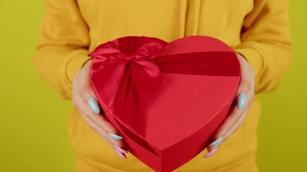 Parte del cuerpo de la mujer con caja de regalo roja en forma de corazón sobre fondo amarillo. Persona irreconocible sosteniendo un regalo romántico con cinta. Concepto de presente en el día de San Valentín y el Día Internacional de la Mujer. — Vídeo de stock