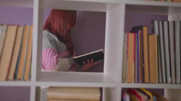 Student čte knihu v knihovně za knihovnami. Zamyšlená mladá žena stojící v knihovně mezi knihami a čtoucí učebnici u fialové zdi