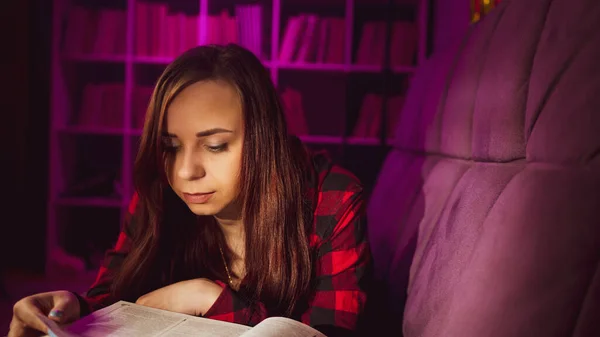 一个漂亮的学生在读一本大书 傍晚时分 一个心事重重的年轻女子躺在沙发上看书 在黑暗的房间里看书 图库图片