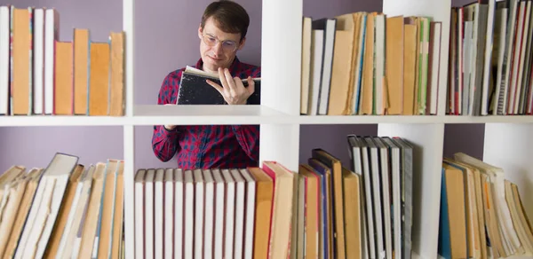 En mand med briller læser en bog i biblioteket bag bogreolerne. En betænksom mand stående i et bibliotek blandt bøger og læser en lærebog mod en lilla væg Royaltyfrie stock-fotos