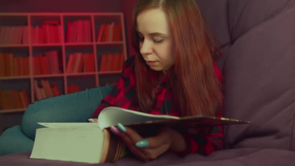 Een mooie student die een groot boek leest. Een attente jonge vrouw liggend op de bank met een boek en lezen in een donkere kamer in de avond — Stockvideo