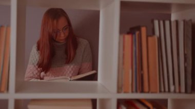 Bir öğrenci kütüphanede kitaplığın arkasında bir kitap okuyor. Kitapların arasında kütüphanede durmuş, mor bir duvara karşı ders kitabı okuyan düşünceli genç bir kadın.
