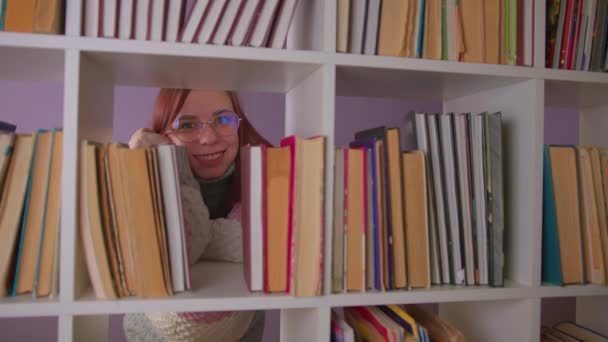 안경을 쓴 젊은 여자는 책꽂이 뒤에 서서 책 과 미소를 통해 카메라를 바라본다. 책장에 의지하여 긍정적 인 감정을 표현하는 행복 한 학생. 도서관에서의 교육 여가 개념. — 비디오