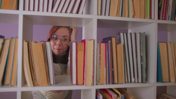 Tänksam kvinna i glasögon tittar på böcker och kliar huvudet, står bakom bokhyllan. Ung kvinna reflekterar över vilken bok man ska läsa. Begreppet stort urval i biblioteket. — Stockvideo