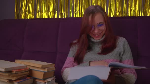 Estudiante cansada leyendo entre libros. Mujer joven y pensativa sentada en el sofá con un montón de libros y libros de texto de lectura — Vídeo de stock
