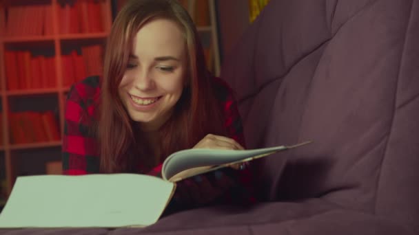 Una hermosa estudiante leyendo un gran libro. Una joven reflexiva tumbada en el sofá con un libro y leyendo en una habitación oscura por la noche — Vídeo de stock